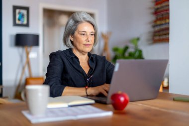 Üst düzey kadın ahşap masada dizüstü bilgisayar kullanarak oturuyor, evden uzaktan çalışıyor, yanında bir elma ve bir fincan kahveyle.