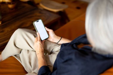 Oturma odasındaki rahat koltukta otururken cep telefonu ve beyaz ekran kullanan yaşlı bir kadın. Makyaj için mükemmel.