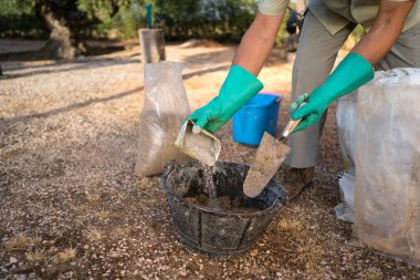 Tanımlanamayan, yeşil eldiven giyen biri çimentoyu bir kürek ve açık havada bir kovayla karıştırıyor. Sahne devam eden bir DIY projesini yakalıyor.