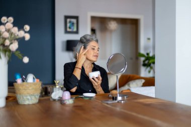 Olgun bir kadın aynaya bakar ve yüzüne yaşlanma önleyici krem sürerek evde günlük güzellik gösterisi yapar.