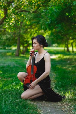 Siyah elbiseli bir kemancı yemyeşil bir parkta diz çöker ve kemanını özenle tutar. Güneş ışığı ağaçların arasından süzülür, dingin ve ilham verici bir sahne yaratır..