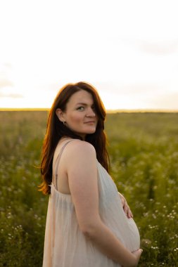 Huzurlu hamile bir kadın güneşli kırsal bir arazide geziniyor ve kameraya bakıyor. Huzurlu an anneliğin güzelliğini ve beklentisini yakalar..