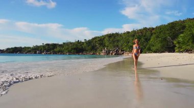 Bir insan sakin bir şekilde Seyşeller sahilinde yürüyor, sakin bir yaz atmosferinin tadını çıkarıyor, berrak mavi sular ve yemyeşil arka planda..