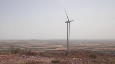 Geniş bir kırsal alanın ortasında, sürdürülebilir ve yeşil enerji çözümlerinin gelişmesini sembolize eden, daha temiz bir geleceğe güç veren yalnız bir rüzgar türbini..
