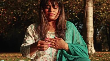 Hintli bir kadın doğal ortamında huzur içinde meditasyon yapıyor, gözleri kapalı, elinde tütsü çubuğu tutuyor. Doğanın sükunetiyle çevrili geleneksel giysiler giyiyor..