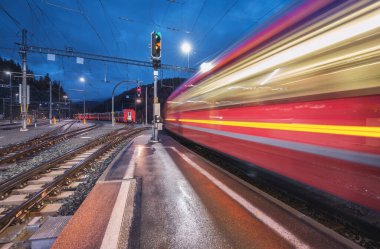 Bulanık kırmızı yolcu treni gece İsviçre Alpleri 'ndeki dağ tren istasyonundan geçiyor. Günbatımında hız treni hareket ediyor. St. Moritz, İsviçre. Bernina Ekspresi. Yağmurda ve ışıklarda demiryolu platformu