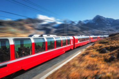 Yüksek hızlı kırmızı tren sonbaharda mavi gökyüzü ile güneşli bir günde İsviçre Alp Dağları 'nda demiryolu boyunca ilerliyor. Bernina Ekspres treni hareket bulanıklığı etkisi, turuncu çimenler, gün batımında karlı kayalar