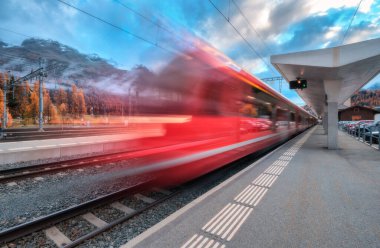 Bulanık kırmızı yolcu treni alacakaranlıkta İsviçre Alpleri 'ndeki dağ tren istasyonundan geçiyor. Hızlı tren hareket ediyor. St. Moritz, İsviçre. Bernina Ekspresi. Demiryolu platformu ve bulutlu gökyüzü. Demiryolu taşımacılığı