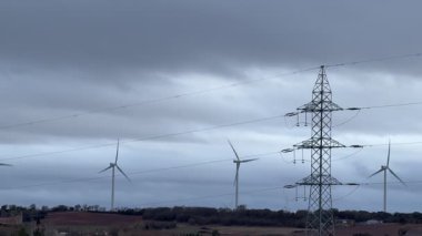 Görüntü, bulutlu bir tepede rüzgar türbinleri sergiliyor, yanında da enerji üretiminde doğa ve teknoloji arasındaki dengeyi gösteren yüksek bir elektrik hattı kulesi var..
