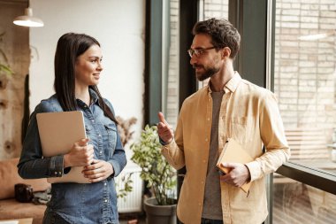 Sakallı adam elinde defterle neşeli bir kadınla konuşurken el kol hareketi yapıyor.