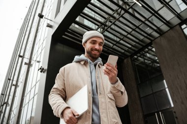 Şapkalı, şapkalı, dizüstü bilgisayarlı ve akıllı telefonlu mutlu serbest çalışanın düşük açılı görüntüsü 