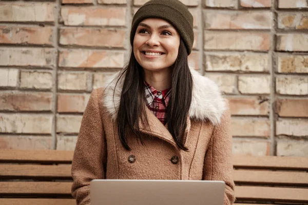 smiling brunette freelancer in beanie hat using laptop while sitting on bench