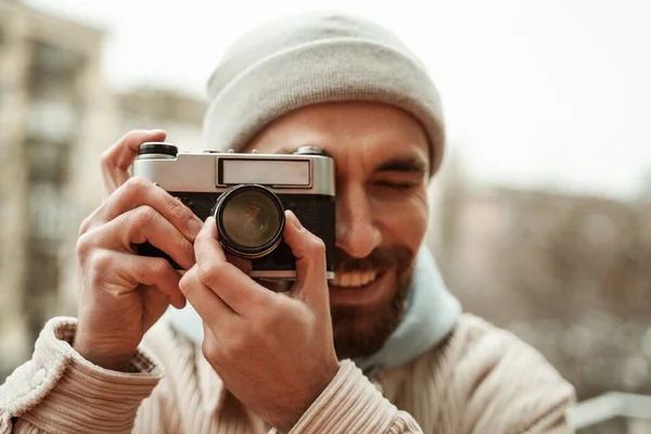 bearded photographer in beanie hat smiling while taking photo on retro camera