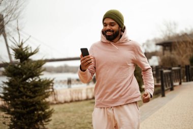 Neşeli Afro-Amerikan koşucu dışarıda koşarken elinde cep telefonu tutuyor.