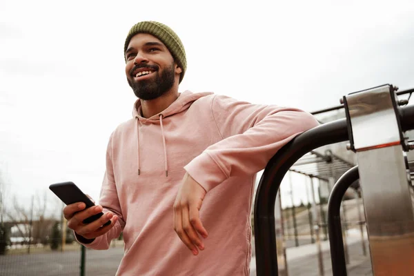 joyful african american sportive man looking away while holding mobile phone outdoors