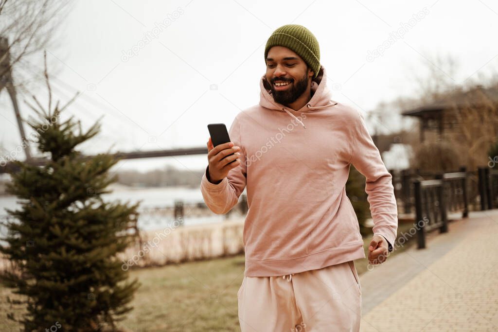 cheerful african american jogger holding mobile phone while running outdoors