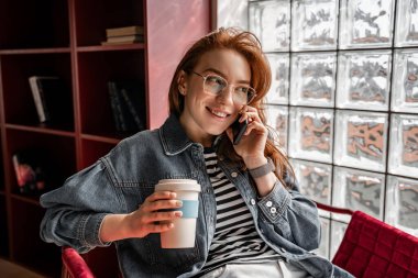 Gözlüklü mutlu kızıl saçlı öğrenci elinde kahve, elinde de akıllı telefonuyla konuşuyor. 