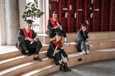 Mezuniyet sınıfı 2021 'de, ellerinde diploma, merdivenlerde oturan mutlu ırklar arası öğrenciler.