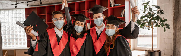multiethnic students in medical masks, graduation gowns and caps holding diploma, banner