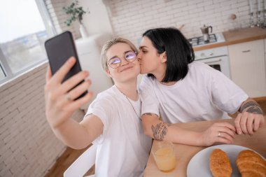 Dövmeli kız arkadaşının yanağını öpen esmer kadın mutfakta selfie çekiyor. 