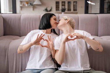 Mutlu lezbiyen çift elleriyle kalp işareti gösteriyor ve oturma odasında öpüşüyorlar. 