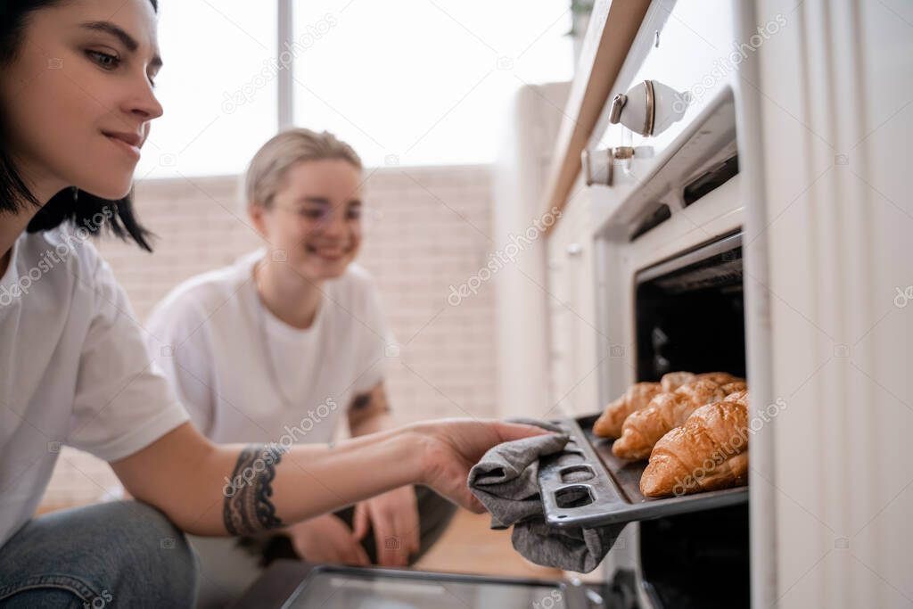 tattooed lesbian couple looking at oven tray with freshly baked croissants in kitchen 