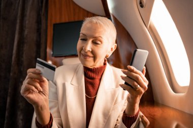 Gülümseyen iş kadını özel jetin kulübesinde kredi kartı ve akıllı telefon tutuyor. 