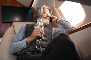 Gülümseyen iş adamının akıllı telefonla konuşması ve uçakta şampanya tutması. 