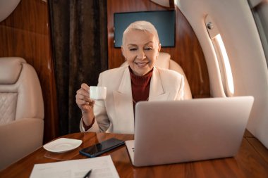 Elinde fincanla gülümseyen iş kadını özel uçaktaki belgenin yanındaki bulanık laptopa bakıyor. 