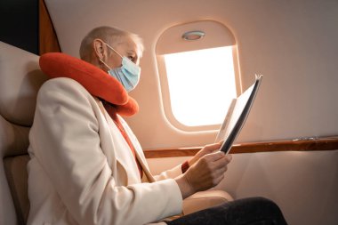 Uçakta medikal maske takan ve boyun yastığı okuyan iş kadını. 