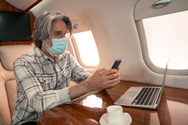 Tıbbi maskeli bir iş adamı laptopun yanında akıllı telefon ve uçakta kahve kullanıyor. 
