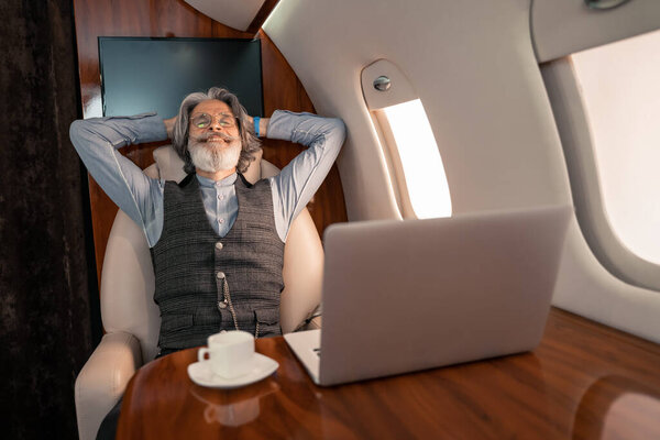 Happy businessman sitting near blurred laptop and coffee in private plane 