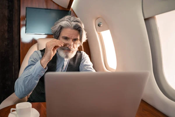 在私人飞机上 在笔记本电脑和咖啡旁边举着眼镜的成熟商人 — 图库照片
