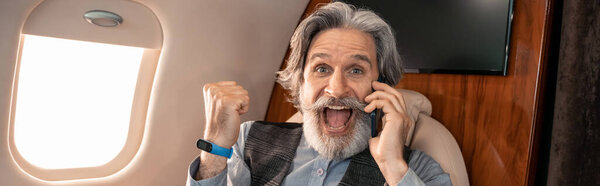 Excited businessman showing yes gesture and talking on smartphone in jet plane, banner 