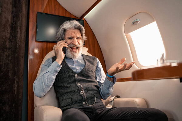 Cheerful mature businessman talking on cellphone in private jet 