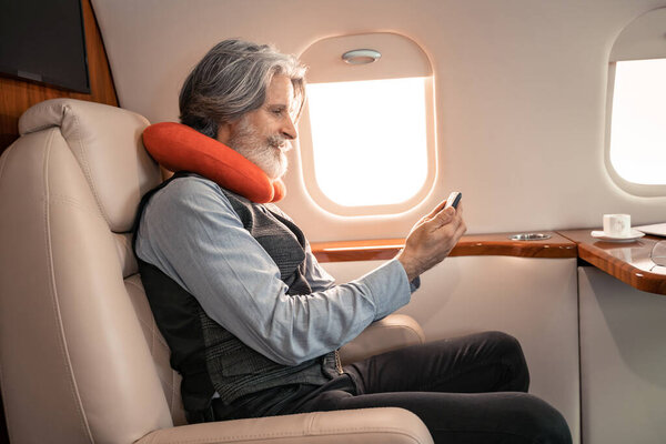 Вид сбоку бизнесмена в шейной подушке с помощью смартфона рядом с кофе в частном самолете 