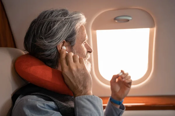 Side view of mature man in neck pillow using earphones in plane