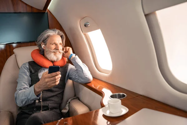 Smiling businessman in earphones and neck pillow holding smartphone in private plane