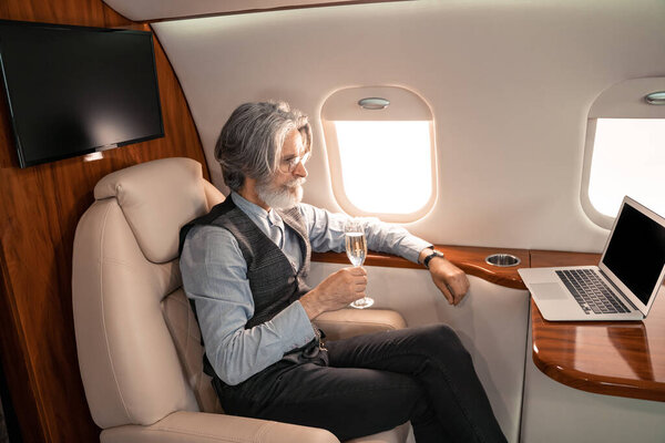 Зрелый бизнесмен с бокалом шампанского смотрит на ноутбук в самолете 