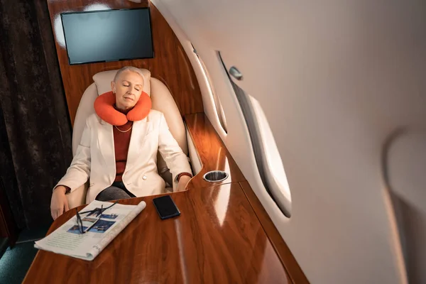 Businesswoman in neck pillow sleeping near magazine and smartphone in private jet