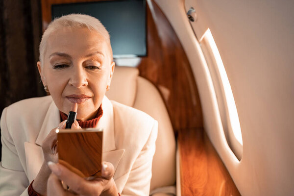 Mature woman holding lipstick and blurred mirror in private plane 