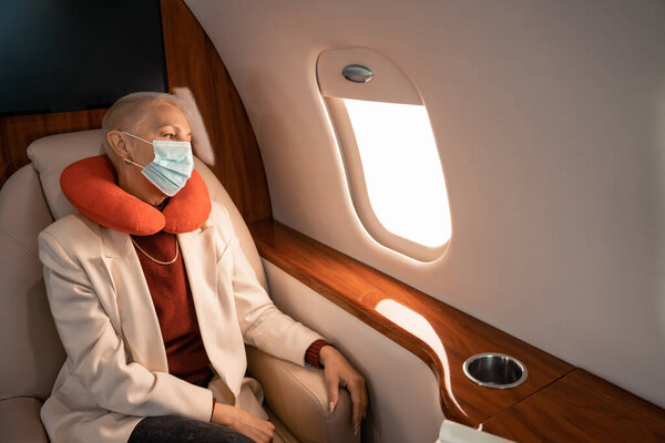 Businesswoman in neck pillow and medical mask looking at plane window 