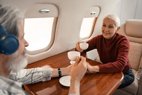 Mujer Sonriente Sosteniendo Taza Café Cerca Marido Borroso Auriculares Avión —  Fotos de Stock