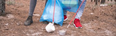 partial view of couple with trash bags picking up rubbish with grabber tools in woods, banner clipart