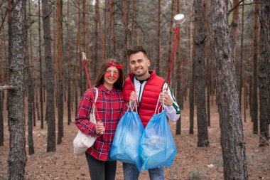 happy couple with trash bags holding pick up tools in forest  clipart
