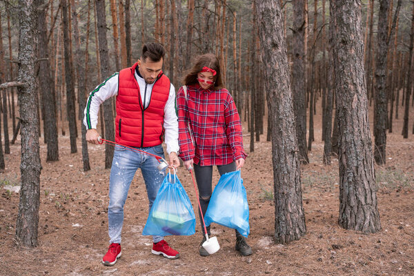 young couple with trash bags picking up rubbish with grabber tools in woods