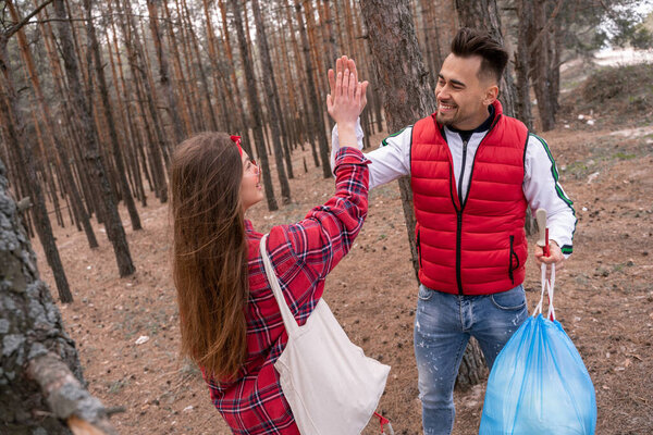 happy man with trash bag giving high five to woman while looking at each other in forest