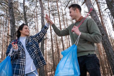 cheerful young woman and man with trash bags giving high five while picking up rubbish with grabbers in forest  clipart