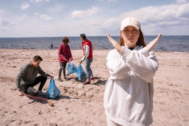 young woman in cap showing stop gesture near group of volunteers picking up trash, ecology concept 