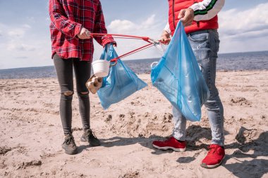 cropped view of couple with trash bags and grabbers picking up garbage on sand  clipart